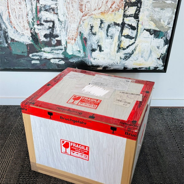 A large cardboard shipping box with red 'FRAGILE' tape and labels sits on a dark textured floor in front of a white wall with an abstract painting.