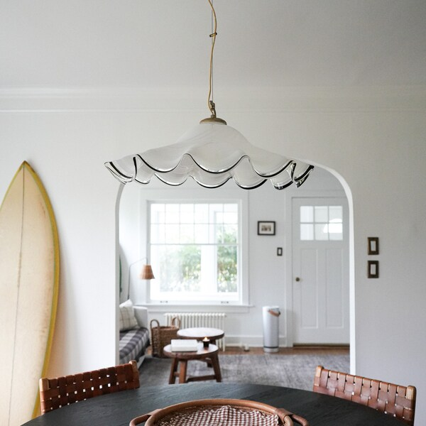 A Murano vintage pendant light with a scalloped white glass shade and black wavy trim hangs in a bright, minimalist dining room. The room features a dark wood table, woven chairs, a surfboard leaning against the wall, and an arched doorway leading to a sunlit living area.